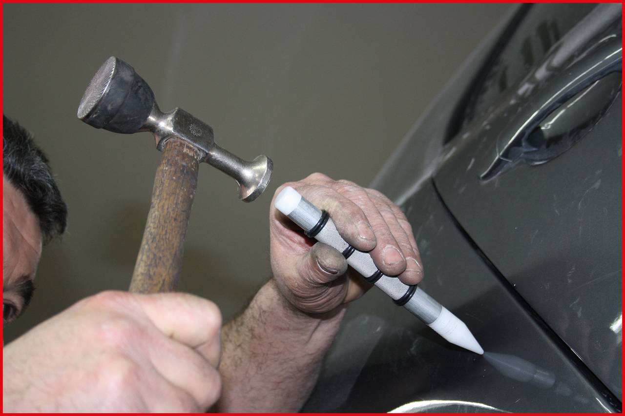 Ein Mechaniker repariert eine Delle in einem grauen Autoteil mit einem Spezialwerkzeug und einem kleinen Hammer.