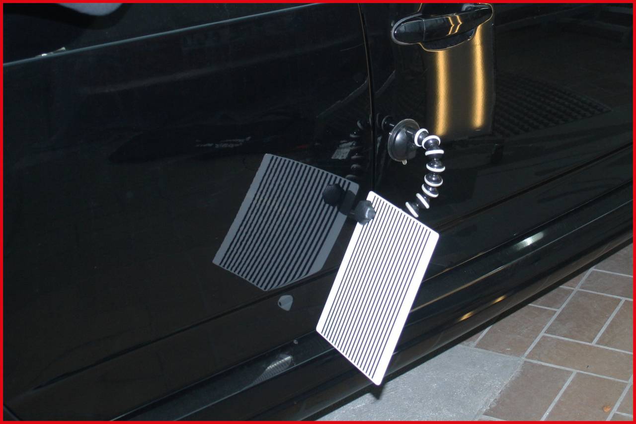 Ein dentales Gerät zur Erkennung von Dellen an einem schwarzen Autotür, bestehend aus reflektierenden Streifen, montiert auf einem flexiblen Arm.