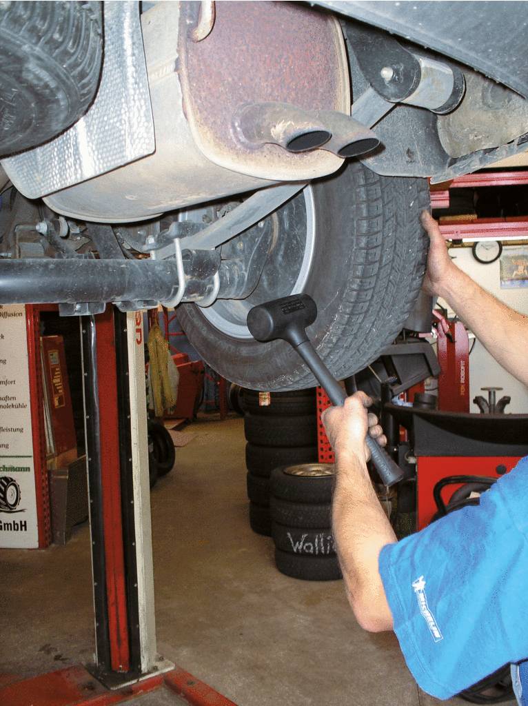 'Person testet Autoreifen in Werkstatt durch Abklopfen mit Gummihammer, erhöhter Wagen auf Hebebühne, Reifen und Auspuff sichtbar.'
