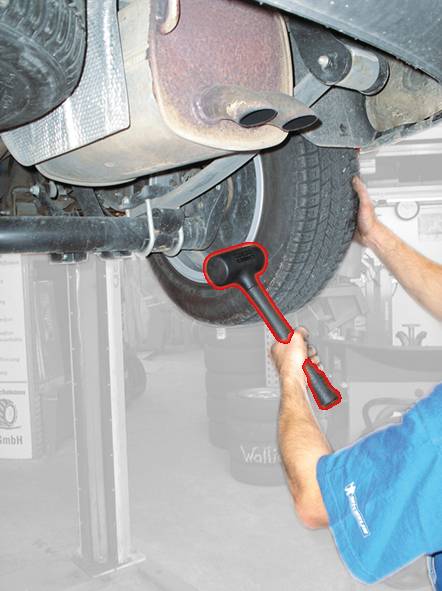 Eine Person repariert ein Auto; sie arbeitet an einem Hinterrad mit einem Hammer, das Auto ist auf einer Hebebühne angehoben.