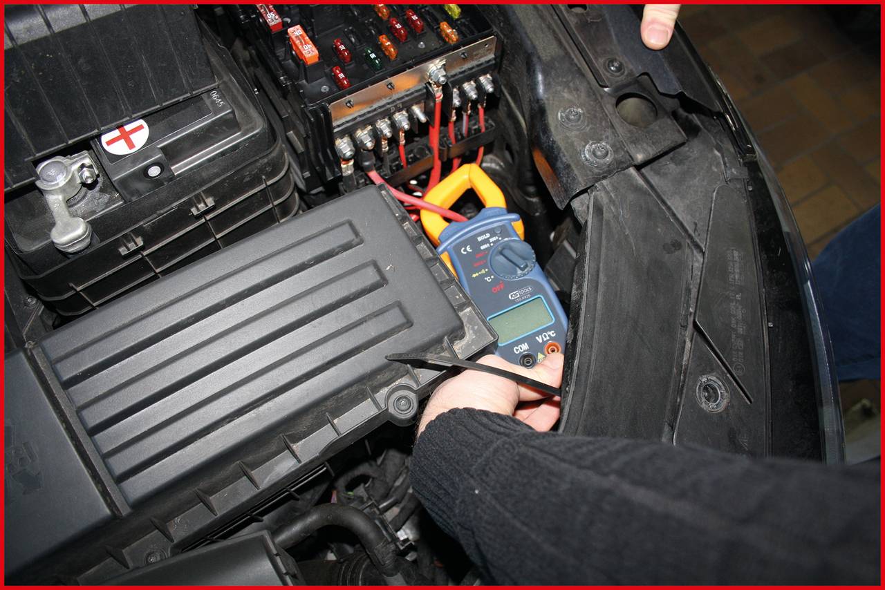 Person misst die Spannung einer Autobatterie mit einem Multimeter. Die Batterie ist in einem schwarzen Gehäuse montiert.