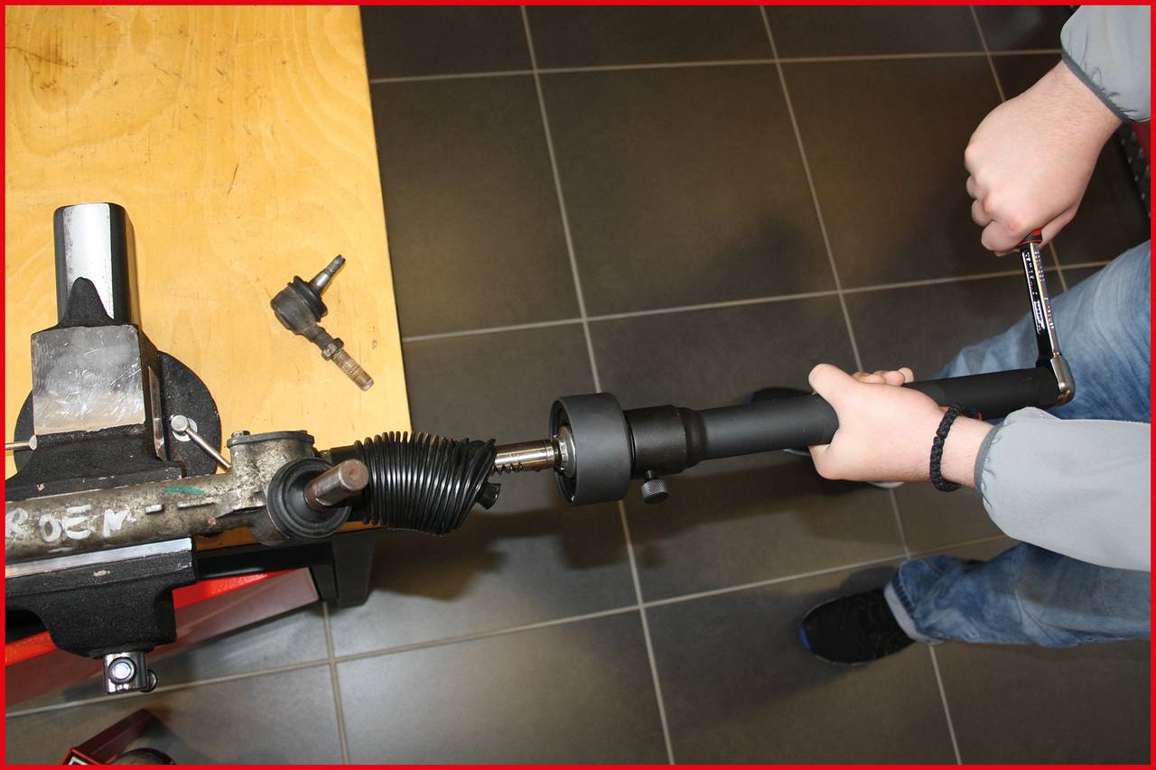 Person repariert eine Auto-Lenkachse in einer Werkstatt. Rechte Hand hält eine Ratsche, linke Hand stabilisiert die Achse.