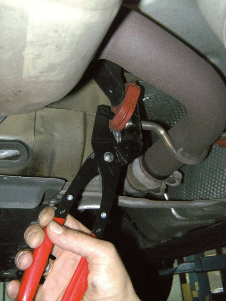 Mechanikerhand hält eine Zange und befestigt eine Metallklemme an einem Auspuff unter einem Fahrzeug.
