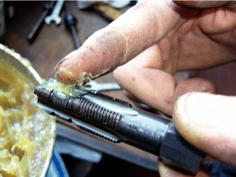 Eine Person trägt mit einem Finger Schmiermittel auf einen metallischen Mechanismus auf. Im Hintergrund sind Werkzeuge sichtbar.