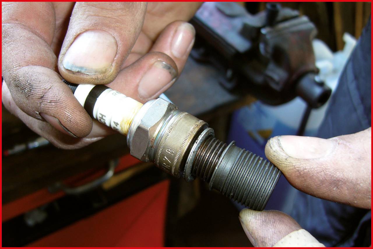 Zwei Hände halten eine Zündkerze, bereit zum Einbau oder Austausch. Im Hintergrund befinden sich unscharfe Werkstattgegenstände.