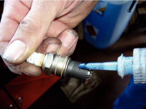 Eine Hand hält eine Zündkerze, während eine andere Hand mit einer Spritze blaue Paste (vermutlich Schmiermittel) aufträgt.