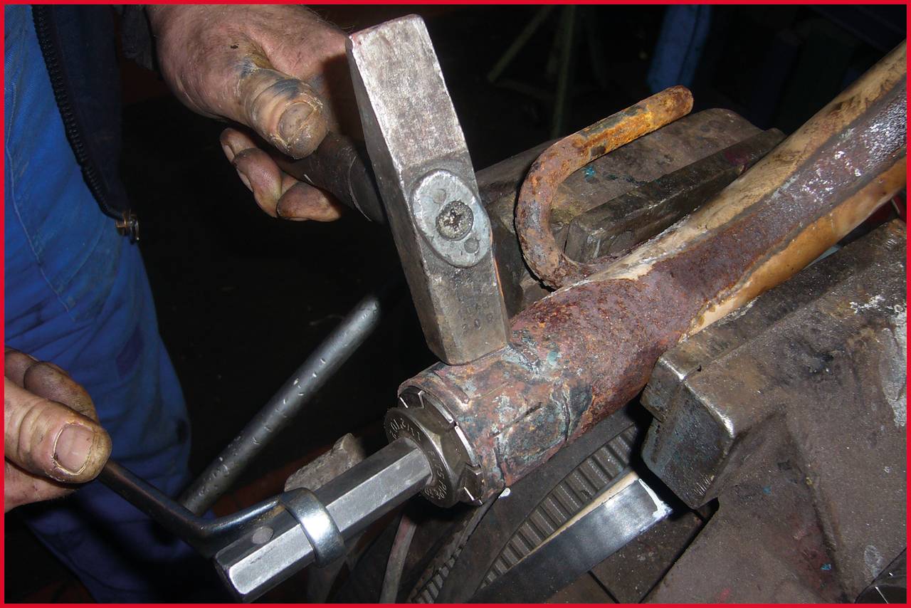 Ein Mechaniker benutzt einen Hammer und einen Schraubenschlüssel, um ein rostiges Rohr in einem Schraubstock zu reparieren. Hände sind im Fokus.