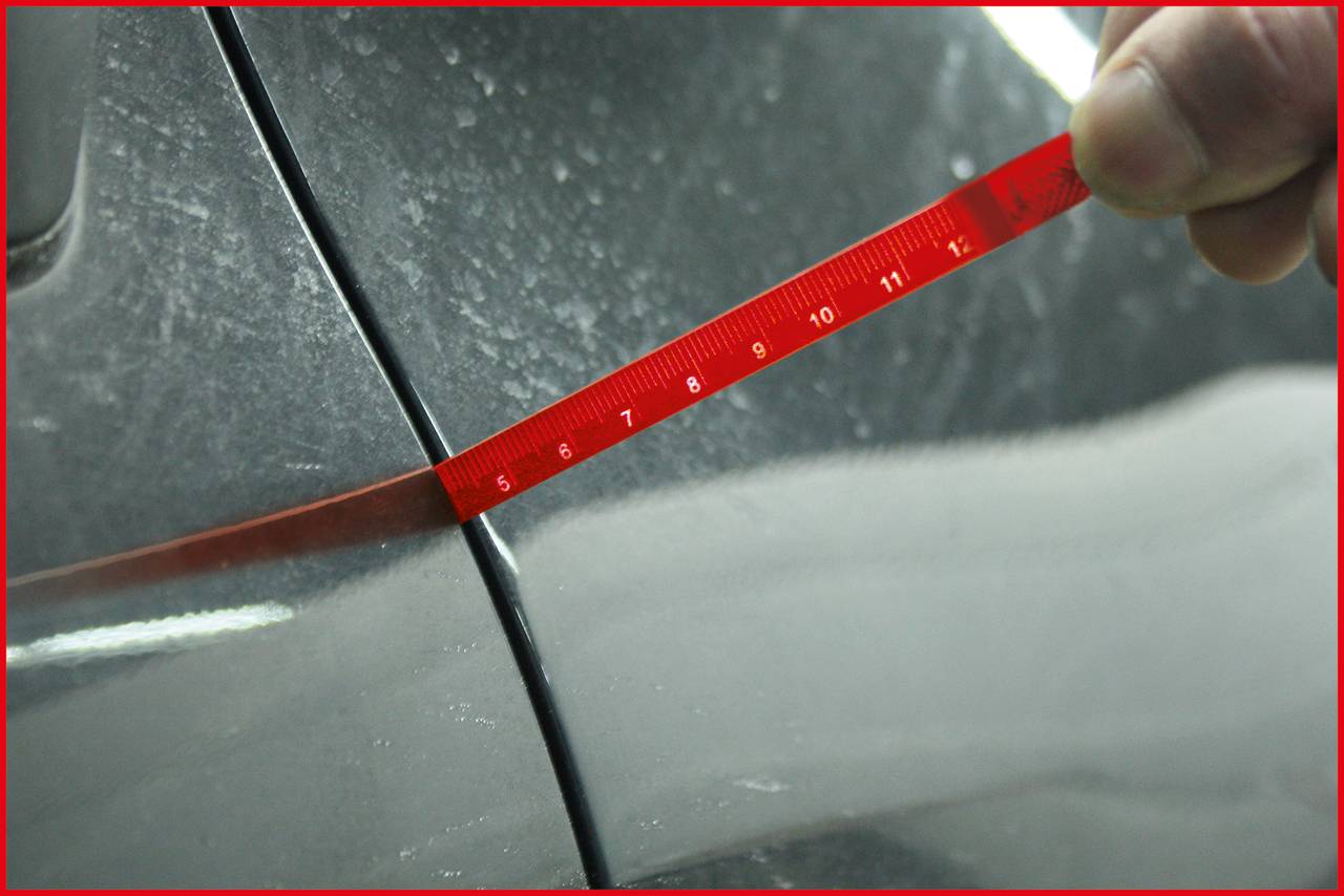 Eine Hand hält ein rotes Lineal an den Spalt zwischen zwei Autoteilen, um den Abstand zu messen.