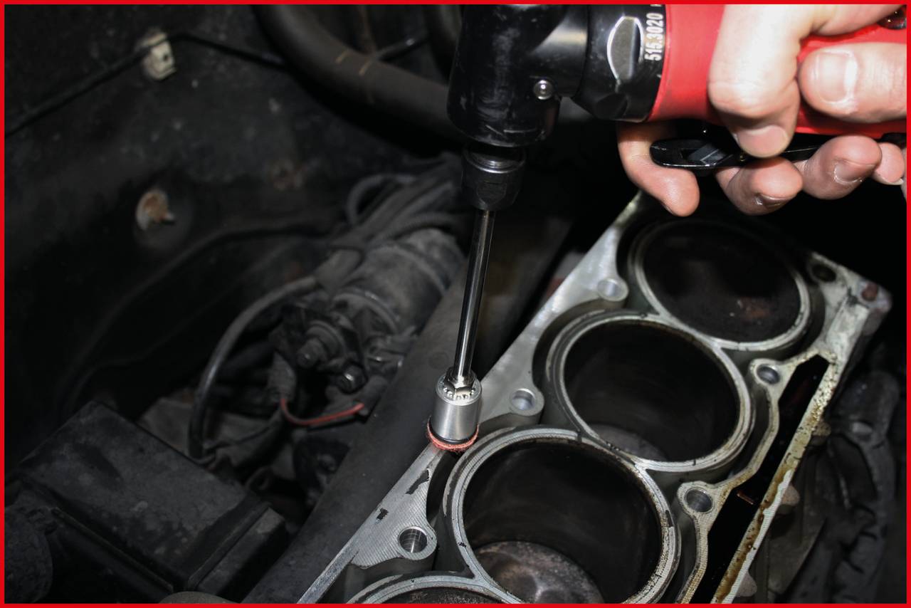 'Eine Hand benutzt einen Elektrowerkzeug, um eine Schraube an einem Zylinderkopf eines Motors anzuziehen. Der Motorraum ist sichtbar.'