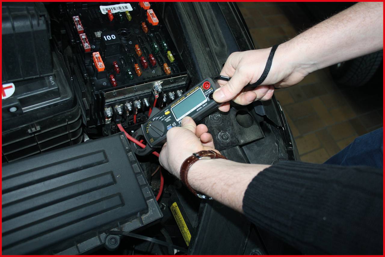 Eine Person misst die Spannung an einer Autobatterie mit einem Multimeter. Im Hintergrund sind Sicherungen und Batteriekabel sichtbar.