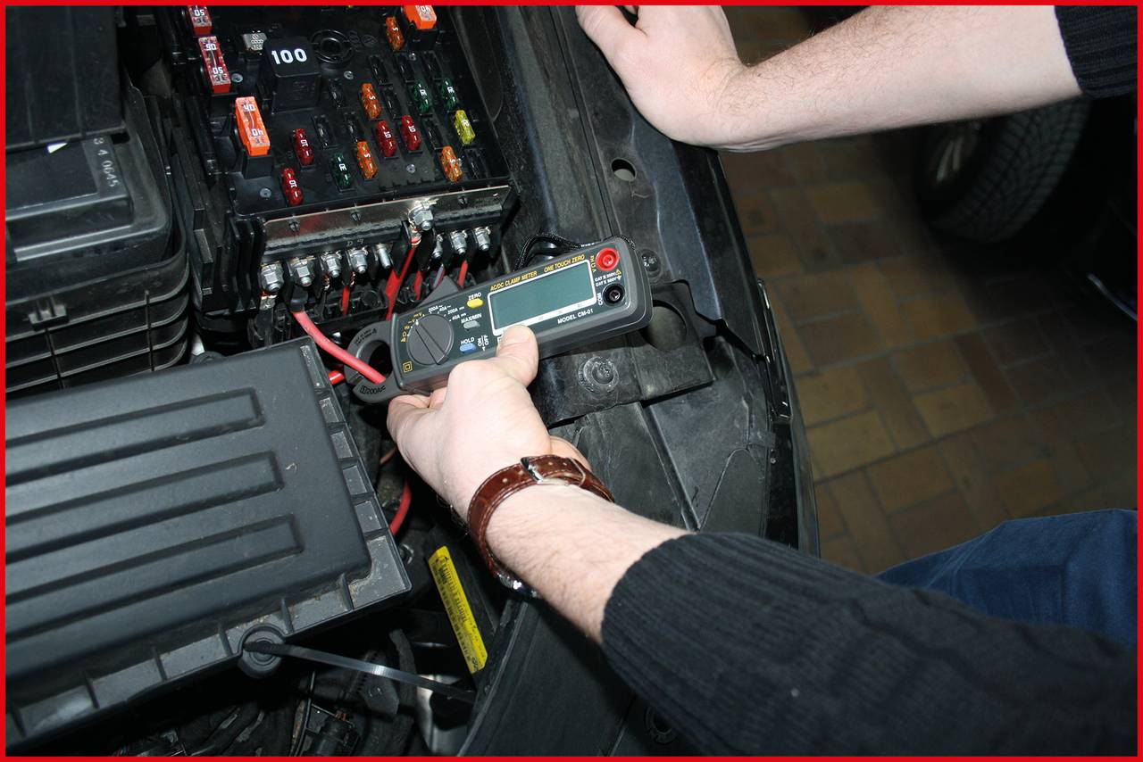 Person misst die Spannung mit einem Multimeter im Motorraum eines Autos. Einige Sicherungen und Kabel sind sichtbar.