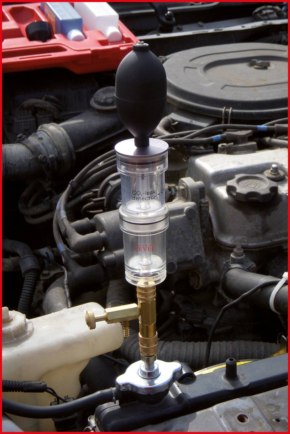 Ein Druckmessgerät ist an einen Autokühler angeschlossen und wird von einer Person daneben kontrolliert. Der Motorraum ist sichtbar.