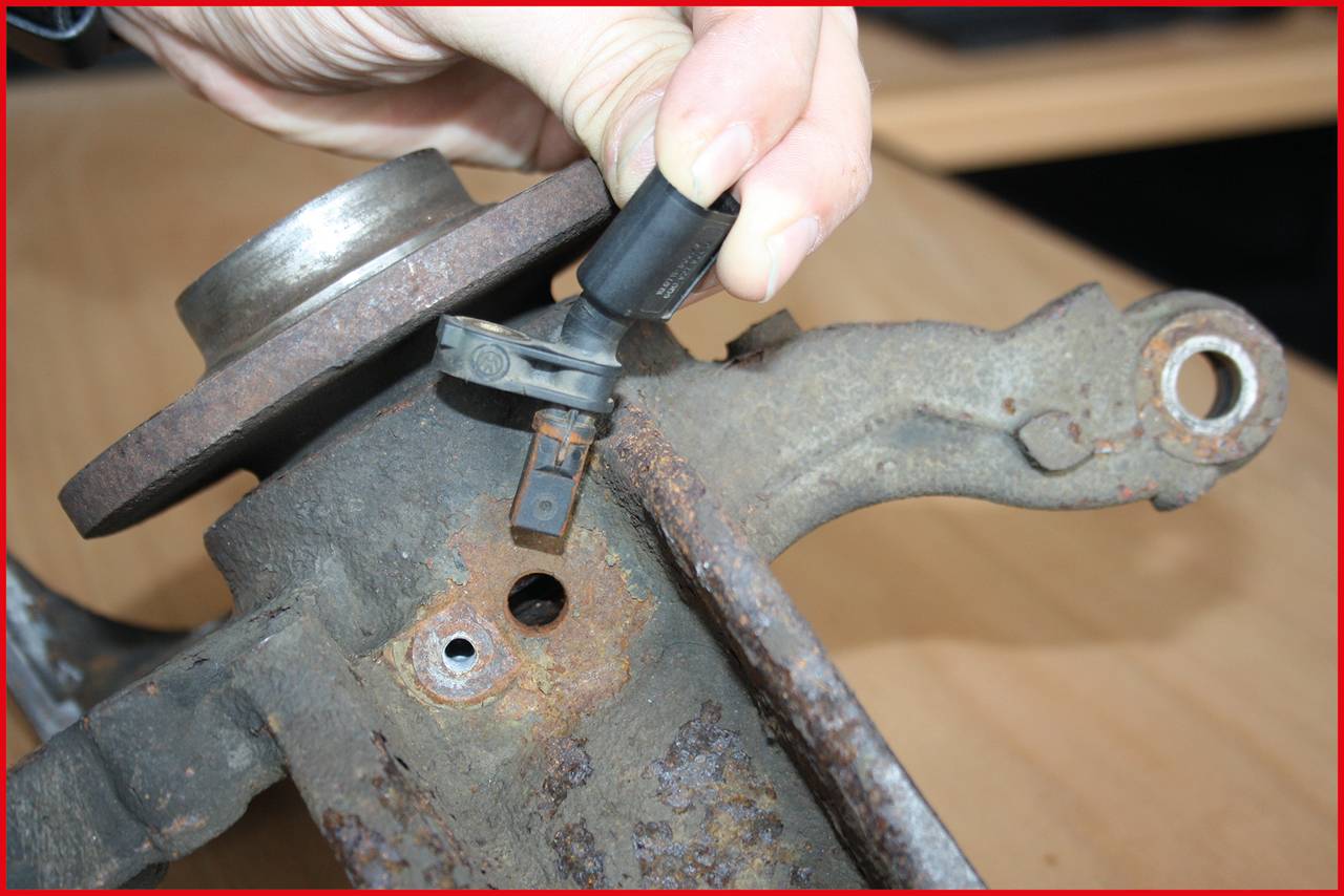 Nahaufnahme einer Person, die einen Sensor direkt an einer rostigen Metallfläche hält, vermutlich ein Teil eines Fahrzeugaufhängungssystems.