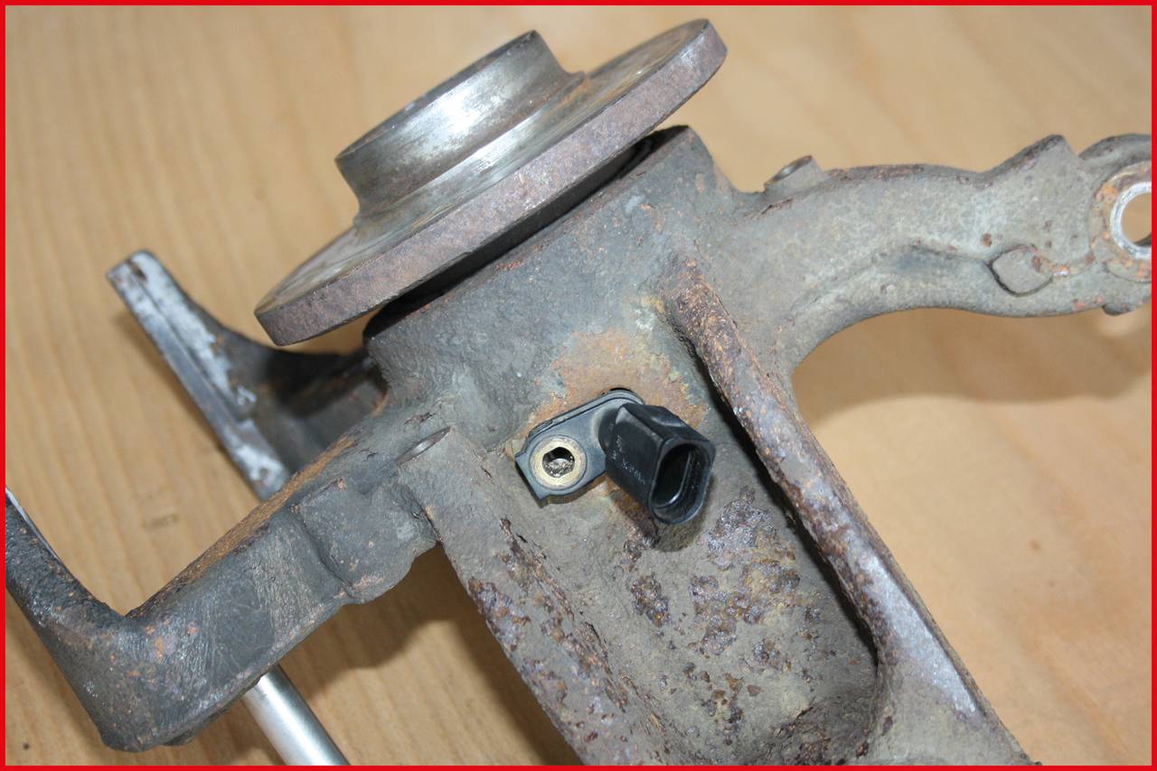Nahaufnahme eines rostigen Autoteils aus Metall auf einer Holzoberfläche, möglicherweise ein Radträger. Ein kleines schwarzes Bauteil ist sichtbar.