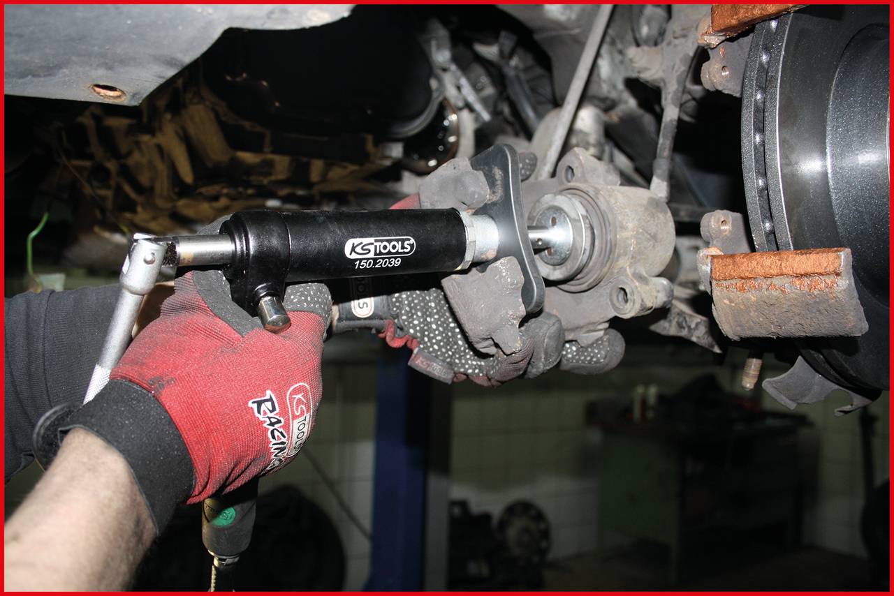 Ein Mechaniker verwendet ein Werkzeug, um an einer Autobremsanlage zu arbeiten. Handschuhe schützen die Hände, während das Fahrzeug angehoben ist.