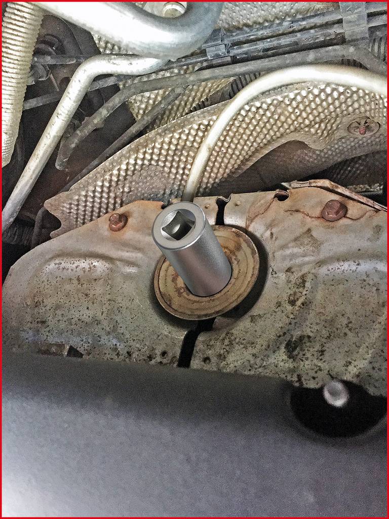 Das Bild zeigt den Unterboden eines Fahrzeugs mit einem schmalen, metallischen Werkzeug, das in einer runden Öffnung befestigt ist.