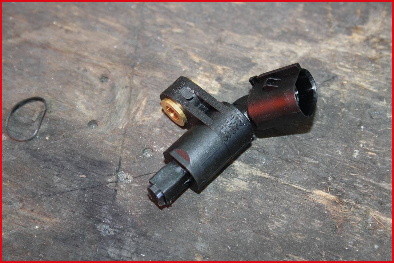 Ein schwarzer Automobil-Sensor liegt auf einem Holztisch. Der Sensor ist zylindrisch mit einem Stecker an einem Ende und einer Befestigungslasche.