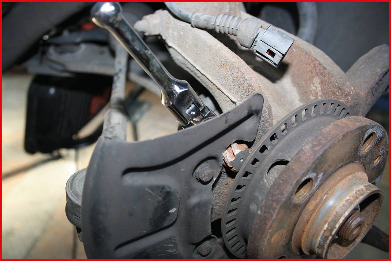 'Nahaufnahme einer Autoreparatur: Eine Ratsche zieht eine Schraube an einer Fahrzeugaufhängung fest. Rostige Bremskomponente im Hintergrund.'