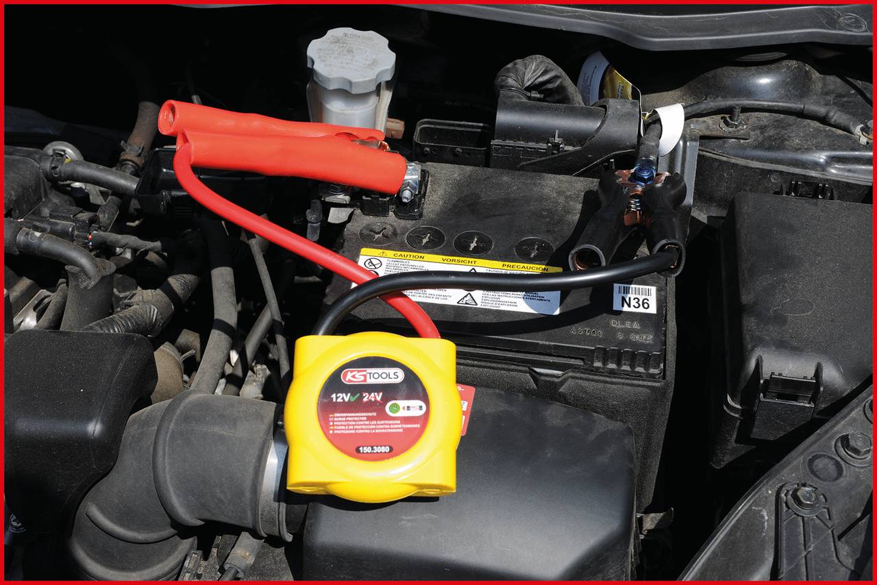 Ein Fahrzeugmotorraum mit angeschlossenen Überbrückungskabeln an der Autobatterie, die rote Klemme ist am Pluspol befestigt.