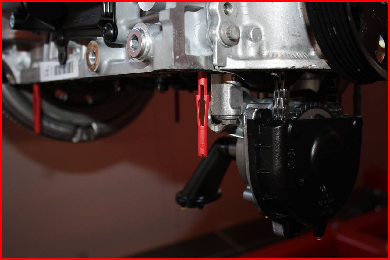 Ein Automotor mit sichtbaren Schrauben und Metallteilen. Im Vordergrund ist ein schwarzes Bauteil und ein rotes Plastikelement erkennbar.