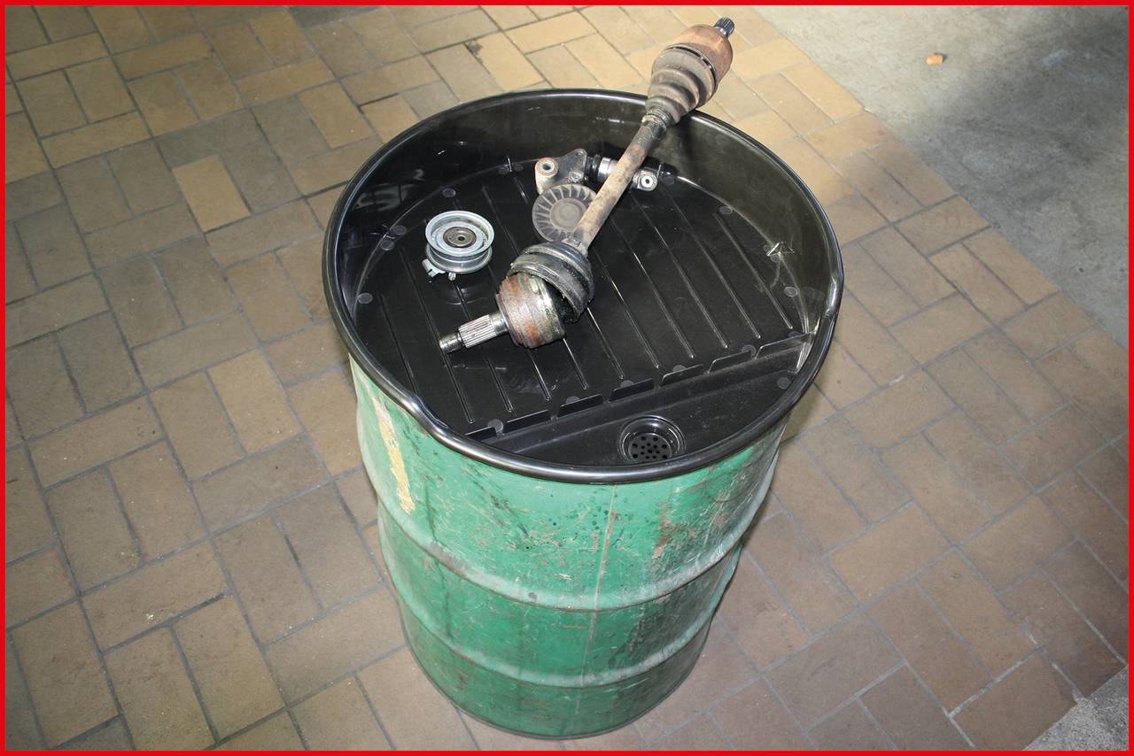 Grünes Metallfass mit schwarzen Deckel steht auf einem Boden aus quadratischen Fliesen. Einige Autoteile liegen obenauf.