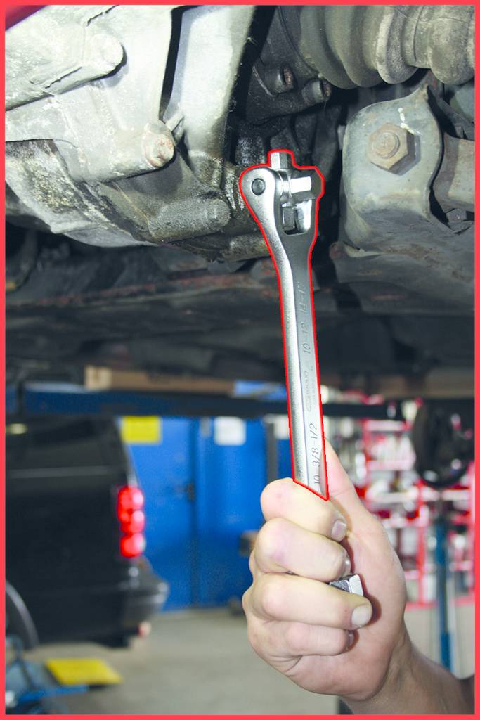 Eine Hand hält einen Schraubenschlüssel und arbeitet an der Unterseite eines Fahrzeugs in einer Werkstatt.