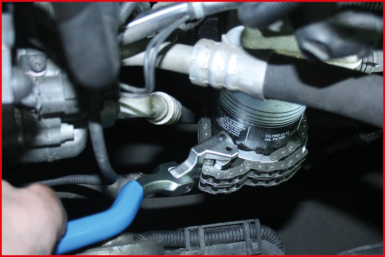 Eine blaue Zange wird verwendet, um einen Ölfilter in einem Automotorraum zu entfernen. Schläuche und Kabel sind im Hintergrund sichtbar.