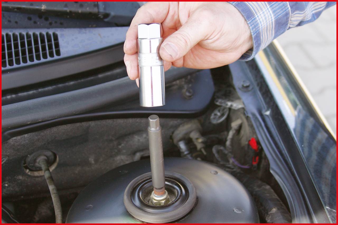 Eine Hand hält eine Stecknuss über dem Federbein in einem geöffneten Motorraum eines Autos, bereit, eine Schraube zu lösen.