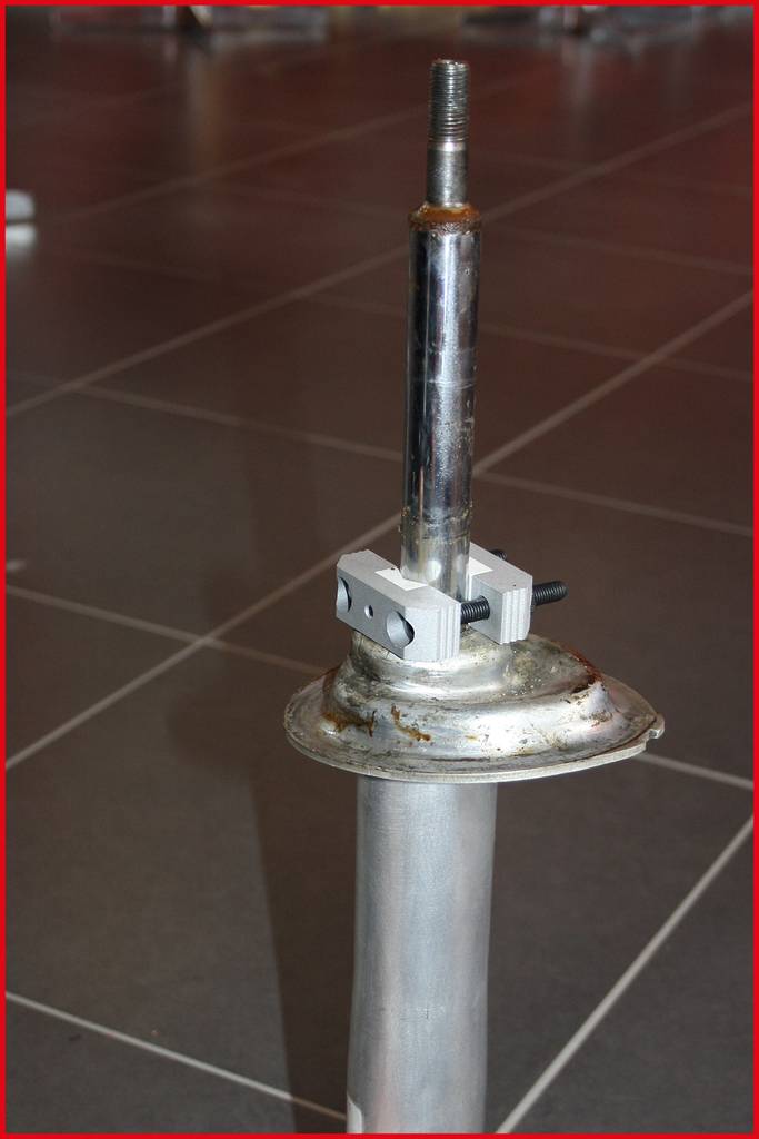 Ein Metallgegenstand mit einem zylindrischen Sockel und einem senkrechten Rohr, das einen drehbaren Kopf mit Schraubmechanismus hält.