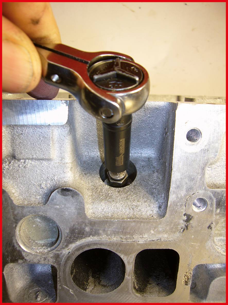 Eine Hand nutzt einen Ratschen-Schlüssel, um eine Schraube auf einem Metallteil festzuziehen. Sichtbar sind einige Bohrungen und Gewinde.