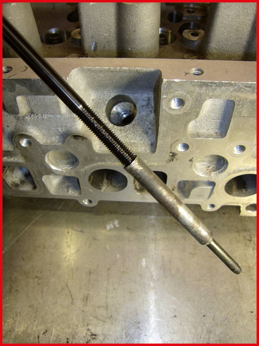 Ein Metallstab mit Gewinde liegt vor einer silbernen, industriell wirkenden Maschine oder Motorblock auf einem Metalltisch.