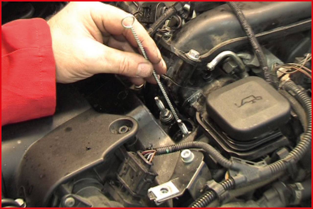 Eine Hand hält eine Zündkerze, die aus einem Auto-Motorraum entfernt wird. Der Motor und Kabel sind im Hintergrund sichtbar.