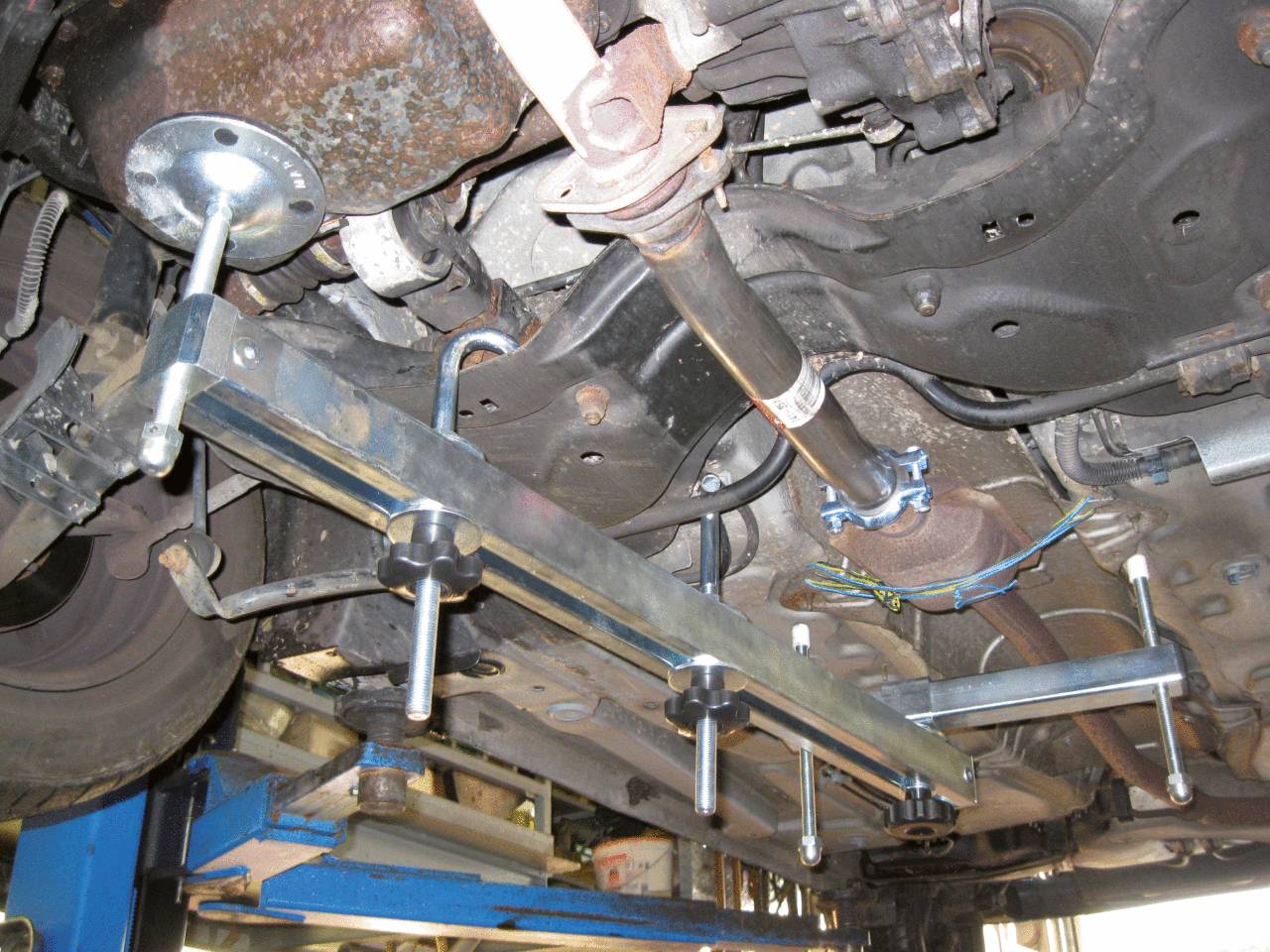 Unterseite eines Autos auf einer Hebebühne. Mechatronische Teile und Gestänge sind sichtbar, möglicherweise im Rahmen einer Reparatur oder Wartung.
