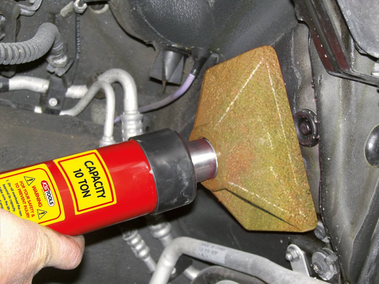 Ein hydraulisches Werkzeug wird verwendet, um die Aufhängung eines Fahrzeugs zu reparieren oder zu justieren. Text auf dem Werkzeug: 'Capacity 10 Ton'.