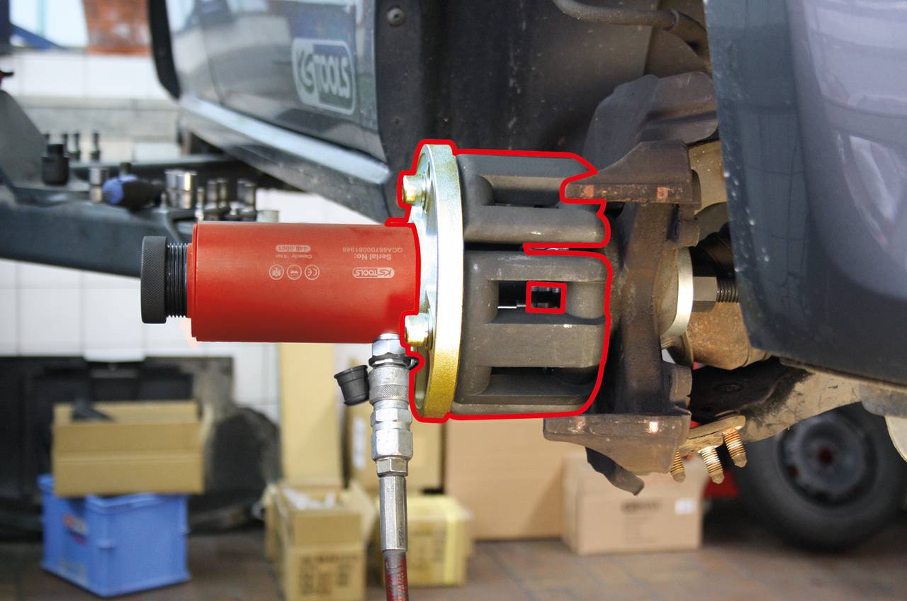 Ein rotes Werkzeug, das an der Radnabe eines Autos montiert ist, um den Radlager-Ausbau zu erleichtern. Im Hintergrund sind Kartons sichtbar.