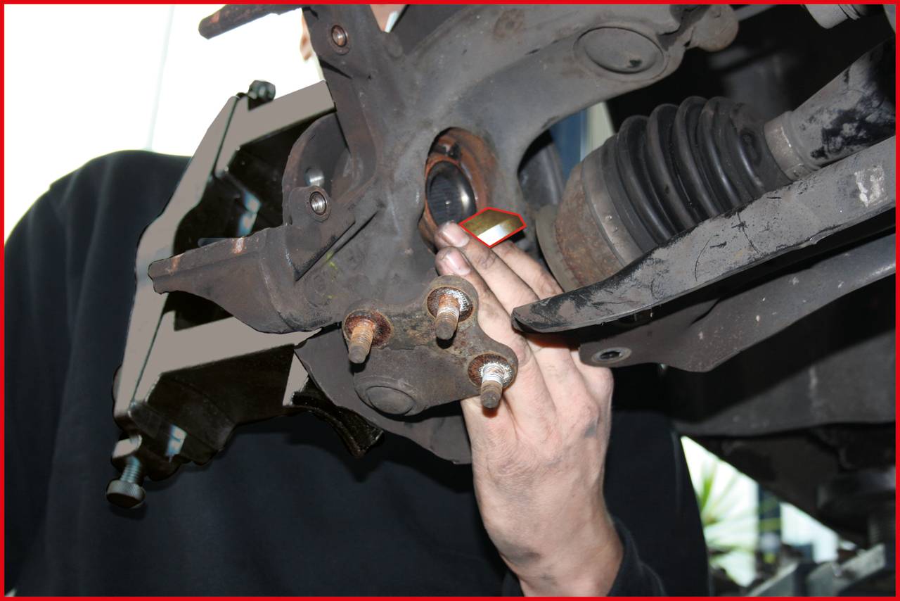 Ein Mechaniker repariert die Radaufhängung eines Autos. Die Hand justiert oder überprüft ein Bauteil. Werkzeuge und Fahrzeugteile sind sichtbar.