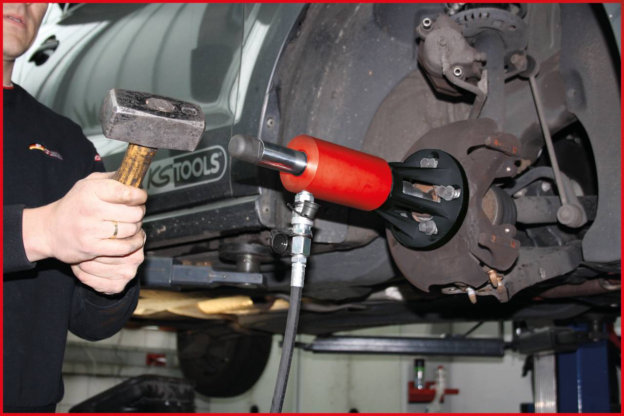 Mechaniker benutzt Spezialwerkzeug und Hammer, um Autoteil an einem Fahrzeug zu reparieren. Vordergrund zeigt Reifenmontagebereich.