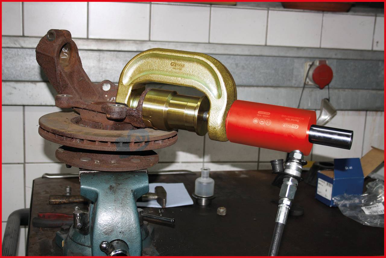 'Bild eines rostigen Bremssattels in einer Werkstattklammer. Ein hydraulisches Werkzeug klemmt den Bremssattel, gezeigt auf einer Werkbank.'