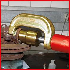 Hydraulischer Abzieher zieht eine alte, rostige Radnabe von einer Achse. Das Werkzeug ist golden und rot und wird in einer Werkstatt verwendet.