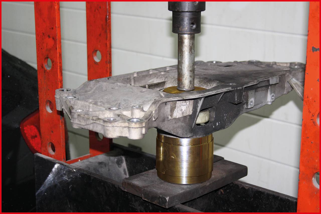 Ein Metallteil wird von einer hydraulischen Presse bearbeitet. Die Presse drückt das Teil auf einen zylindrischen Block. Werkstattumgebung.