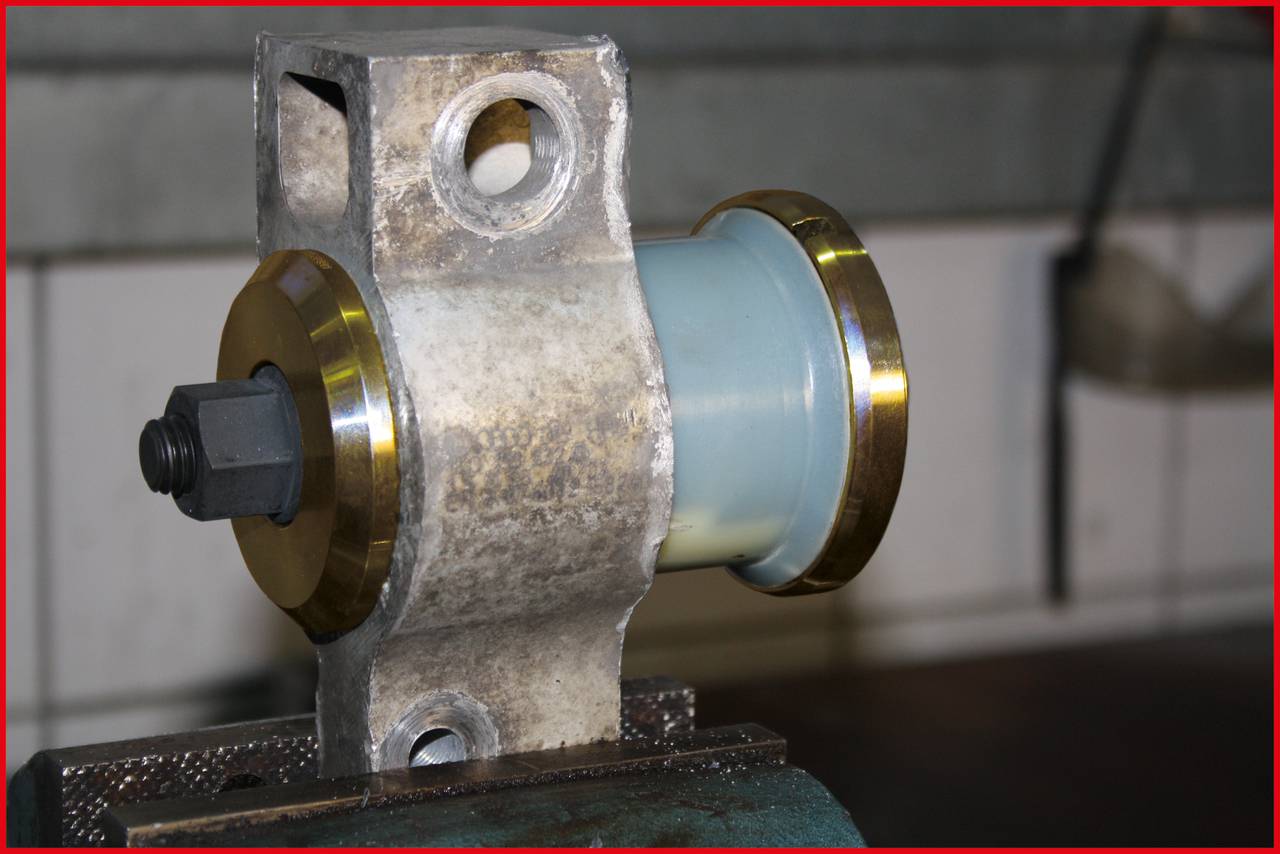 Eine metallische Halterung mit einer zentralen, zylindrischen Rolle, befestigt auf einem Schraubstock in einer Werkstattumgebung.