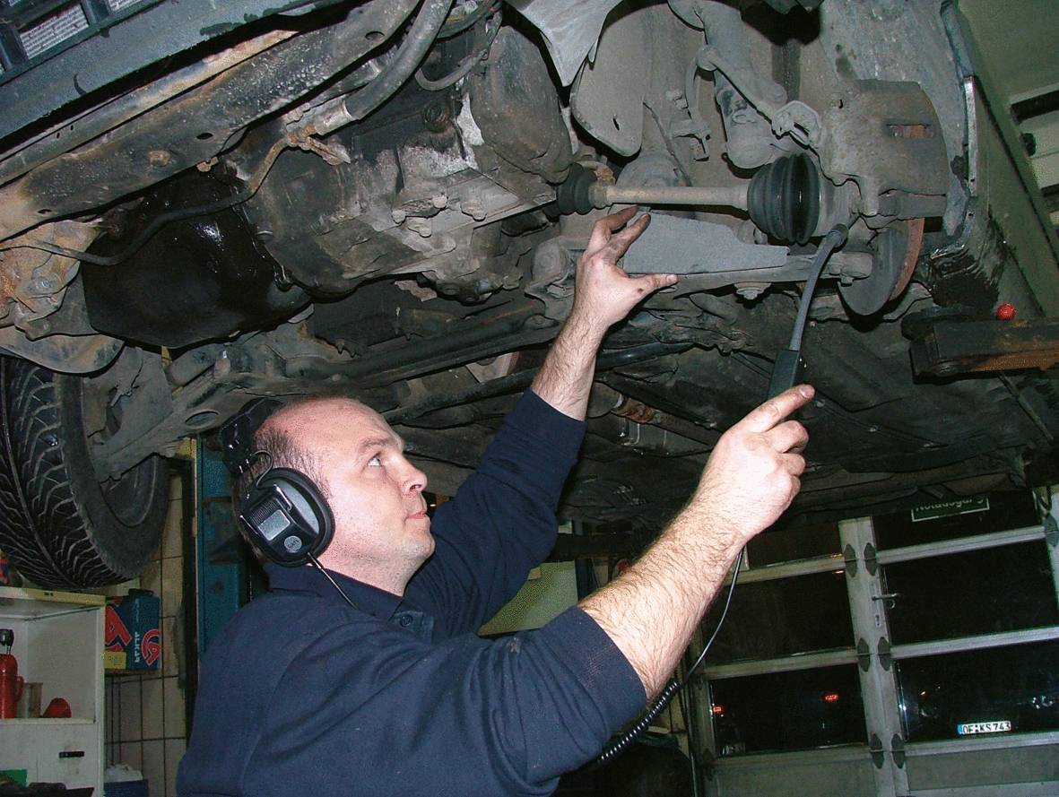 Mechaniker inspiziert das Auto von unten in einer Werkstatt, dabei trägt er Kopfhörer und hält eine Inspektionslampe.