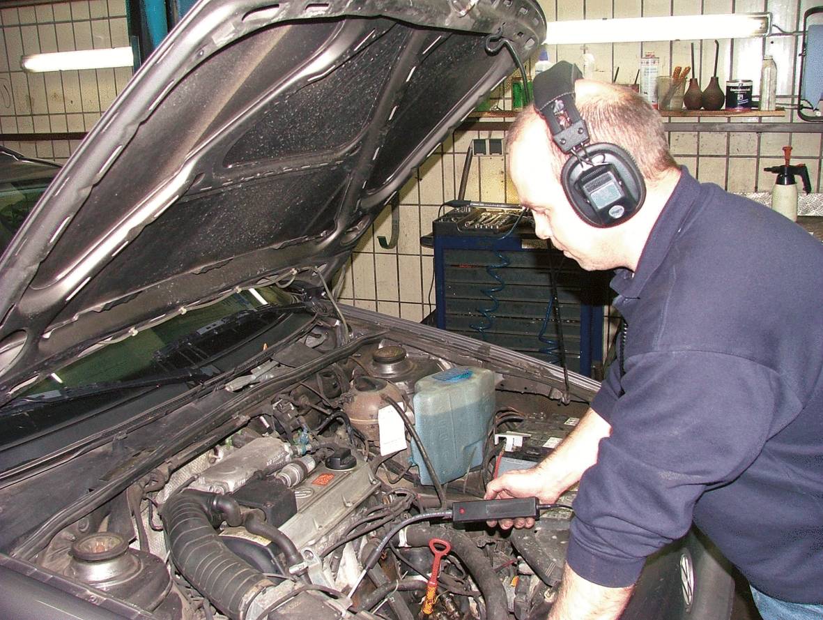Ein Mechaniker mit Kopfhörern überprüft den Motor eines Autos in einer Werkstatt. Die Motorhaube ist geöffnet.