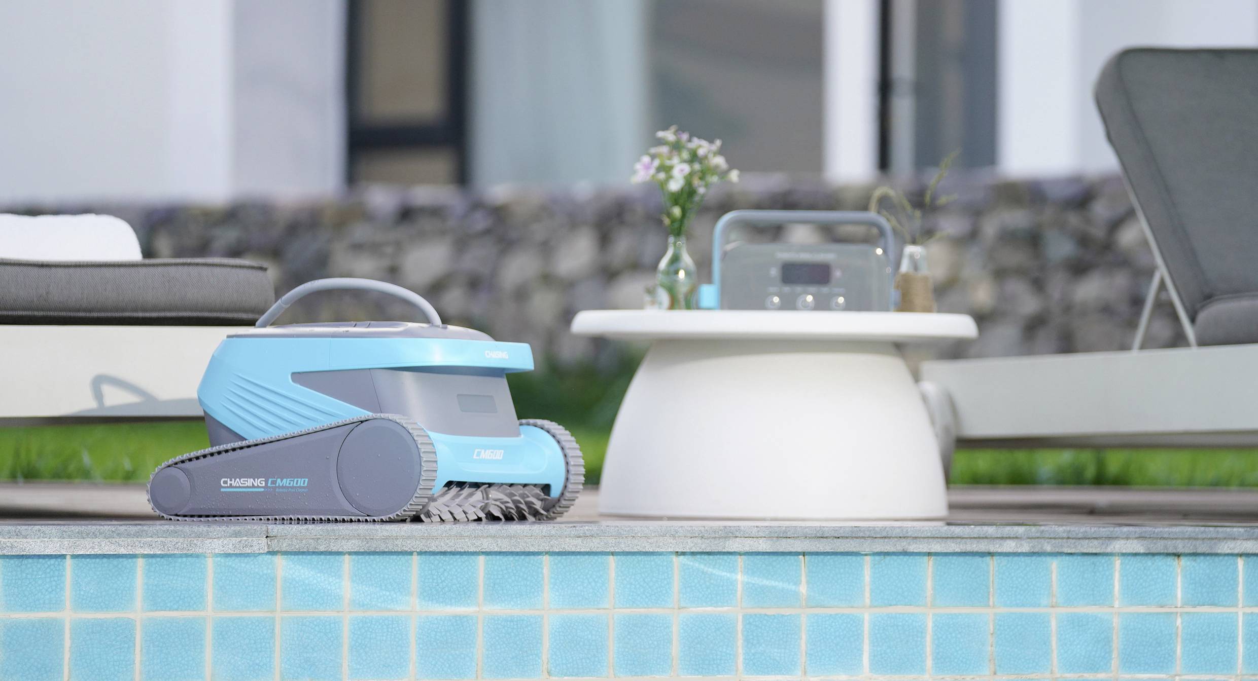 Ein blauer Poolroboter steht am Rand eines Pools neben einem kleinen Tisch mit Radio und Blumen. Im Hintergrund sind Liegestühle sichtbar.