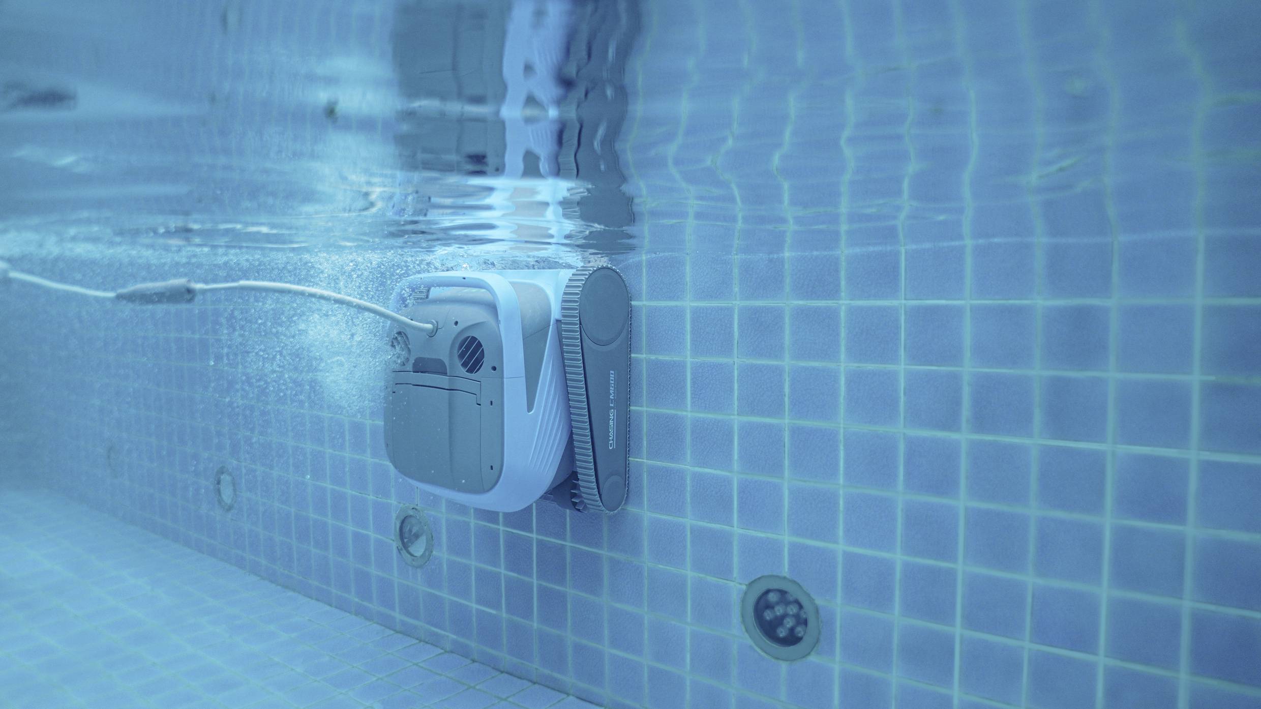 Ein automatischer Poolreiniger reinigt die Wand eines Schwimmbeckens unter Wasser. Er ist rechteckig und mit einem Kabel verbunden.