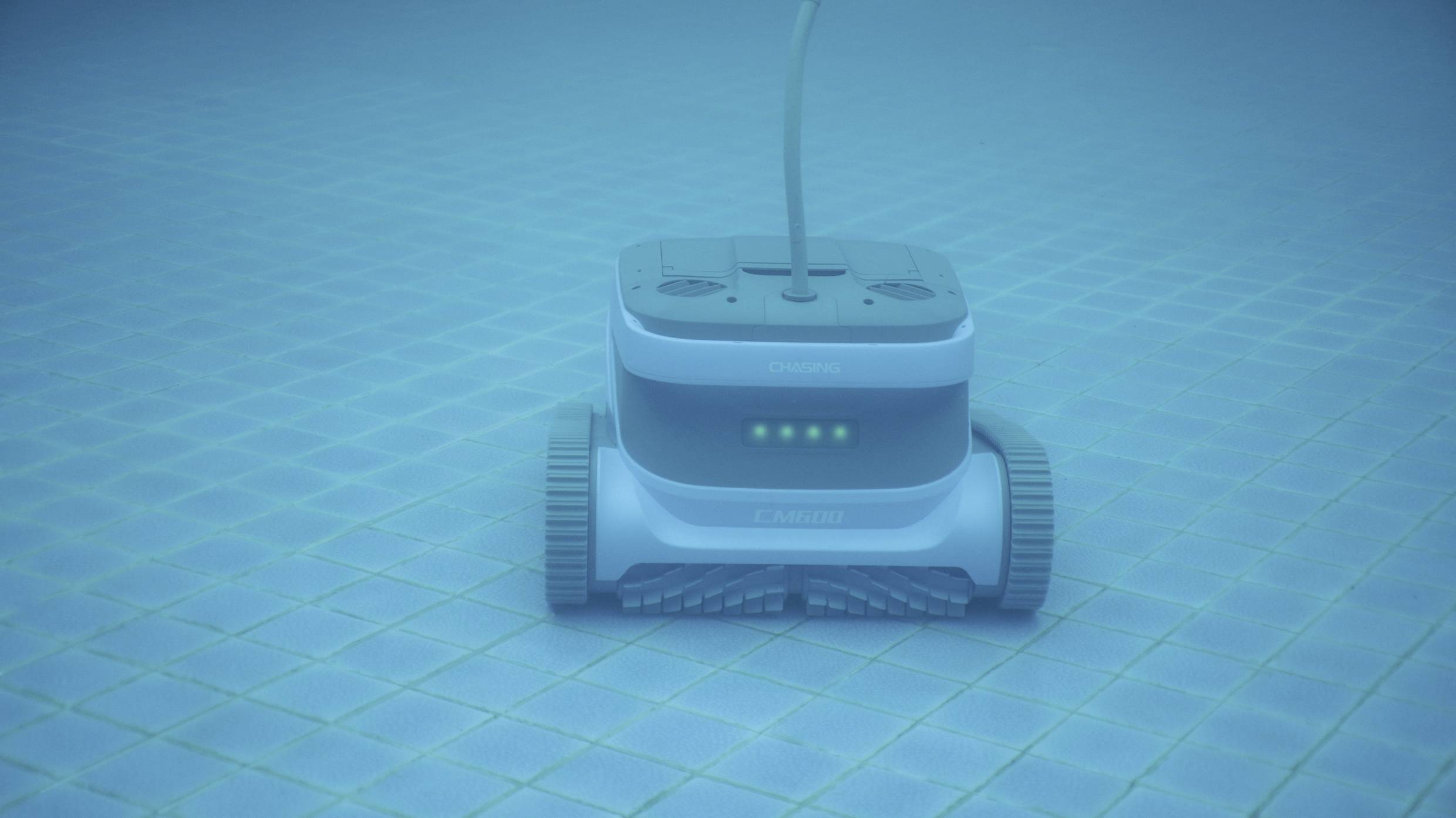 Ein Roboter-Reiniger bewegt sich auf dem Boden eines Swimmingpools, wobei Fliesenmuster und ein Kabel sichtbar sind.