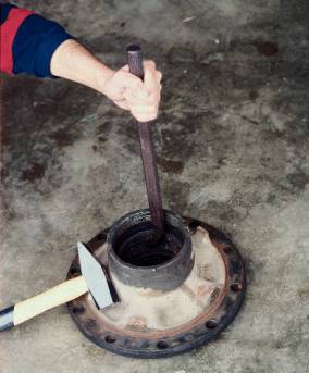 Eine Person bearbeitet ein Metallteil mit einem Hammer und einer Stange auf Betonboden.