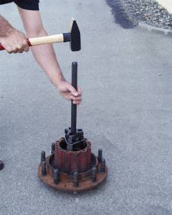 Eine Person hält ein Werkzeug, um einen schweren Metallzylinder auf einem Asphaltboden zu bearbeiten. Das Werkzeug ähnelt einem Hammer.