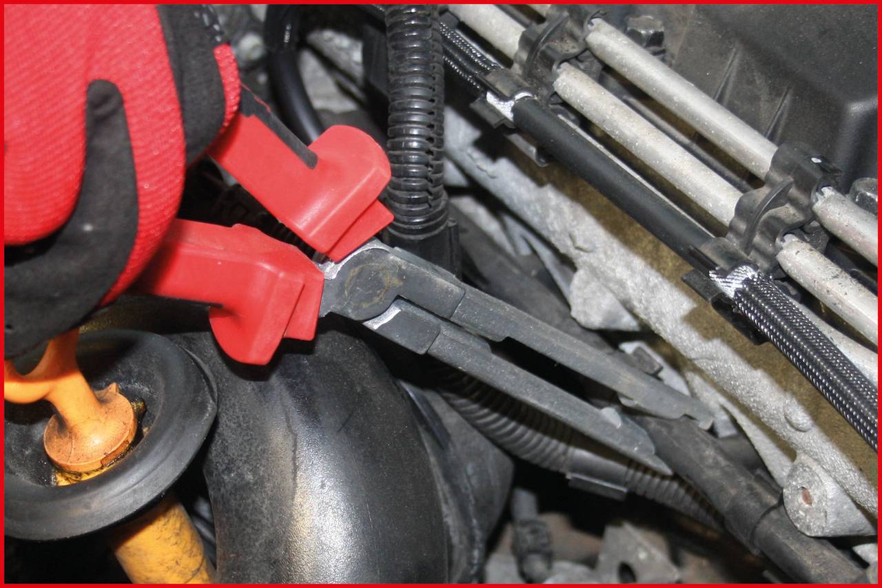Ein Mechaniker arbeitet mit einer roten Zange am Motor eines Fahrzeugs und befestigt oder löst eine Leitung.