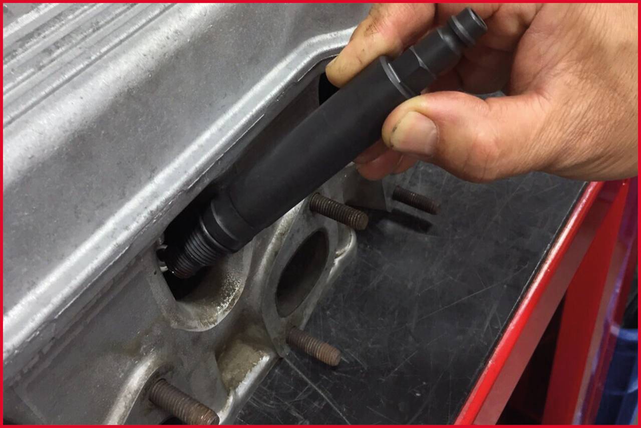 Eine Hand hält eine Zündkerze über einem Motorblock. Die Zündkerze wird zur Installation oder Entfernung positioniert.
